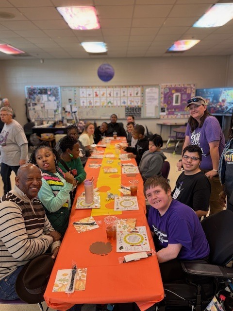 A long table set with orange tablecloths and place settings, decorated for a classroom Thanksgiving feast.