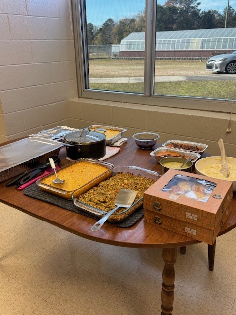 A wooden table buffet with casseroles, stuffing, mashed potatoes, rolls in boxes, and serving utensils by a window.
