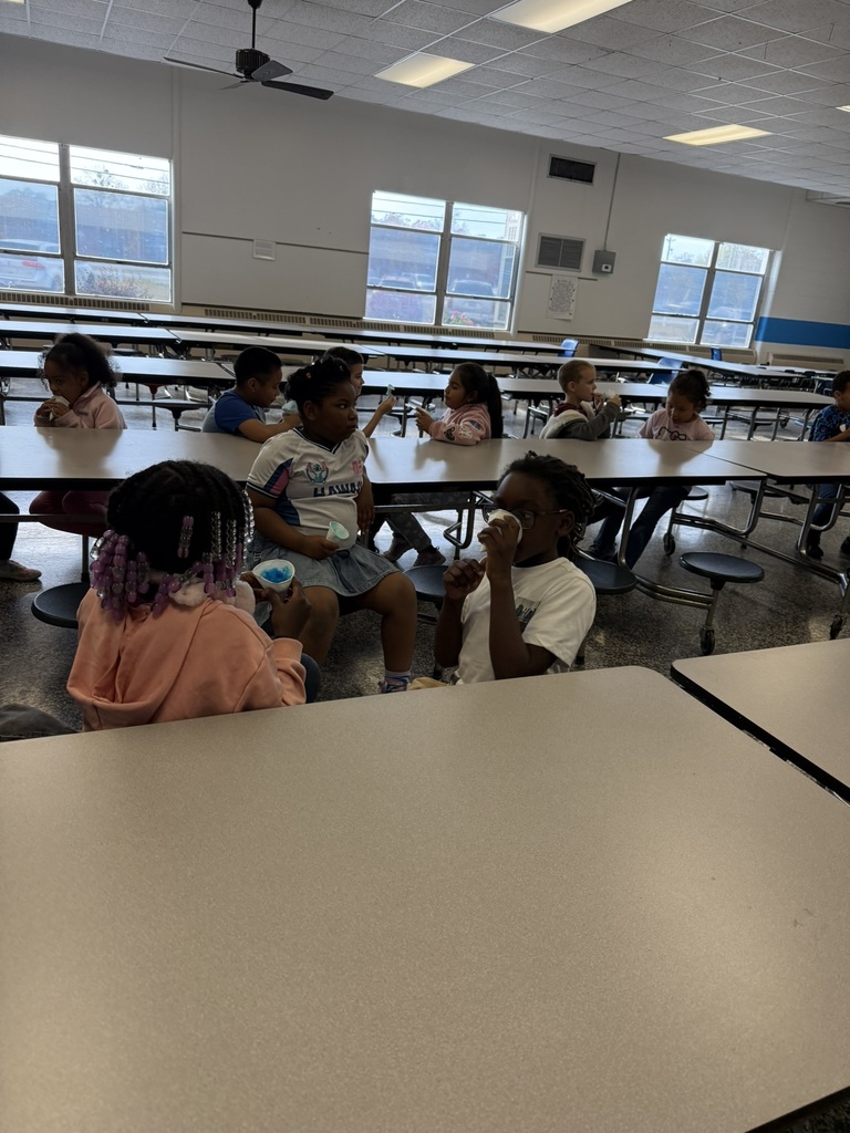 A group of elementary students sit at long cafeteria tables enjoying blue snow cones. Some children chat with friends while others eat quietly. The cafeteria is bright with large windows and empty tables in the background.
