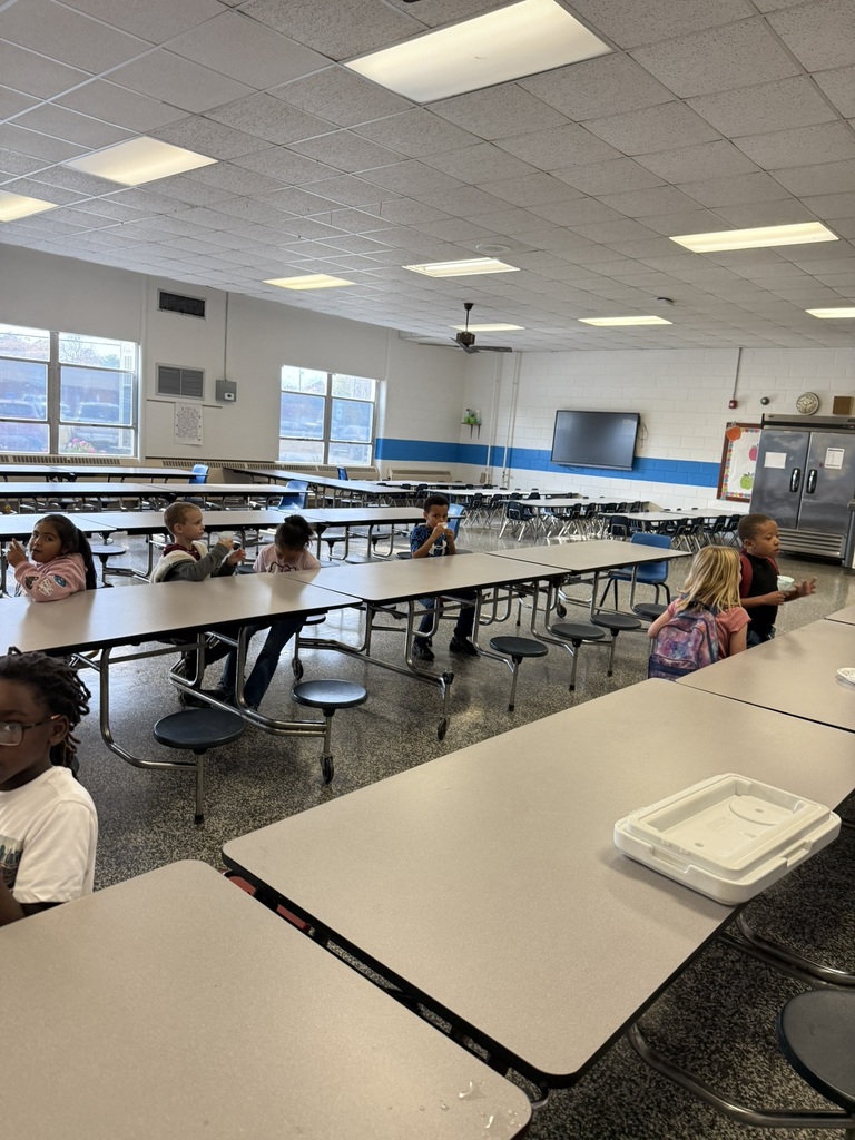 Students sit spread out at cafeteria tables, eating blue snow cones. A few children are chatting, while others look toward the camera. The large cafeteria is mostly empty except for the students participating in the reward activity.