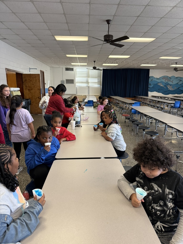 A larger group of children sit at cafeteria tables eating blue snow cones. Several staff members stand nearby helping serve the treats. Students smile and enjoy their snack as part of the good behavior celebration.