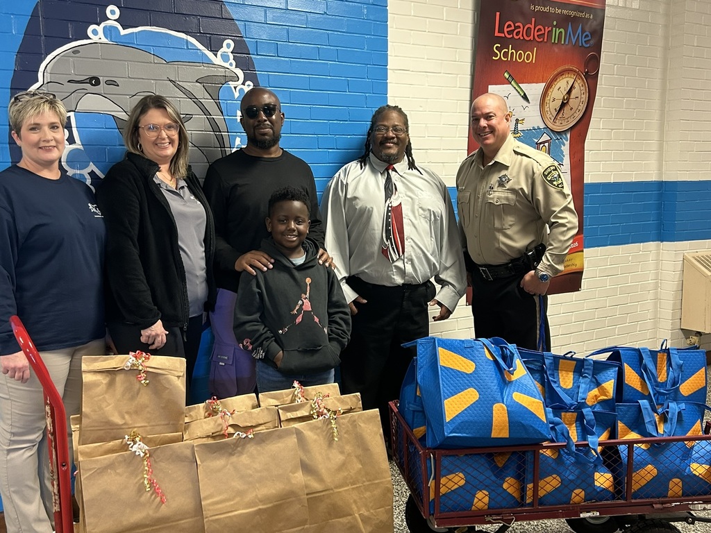 A closer photo of the group smiling in front of the mural and Leader in Me banner. The brown paper bags and blue donation bags are clearly visible in the foreground as part of the Thanksgiving food donation effort.