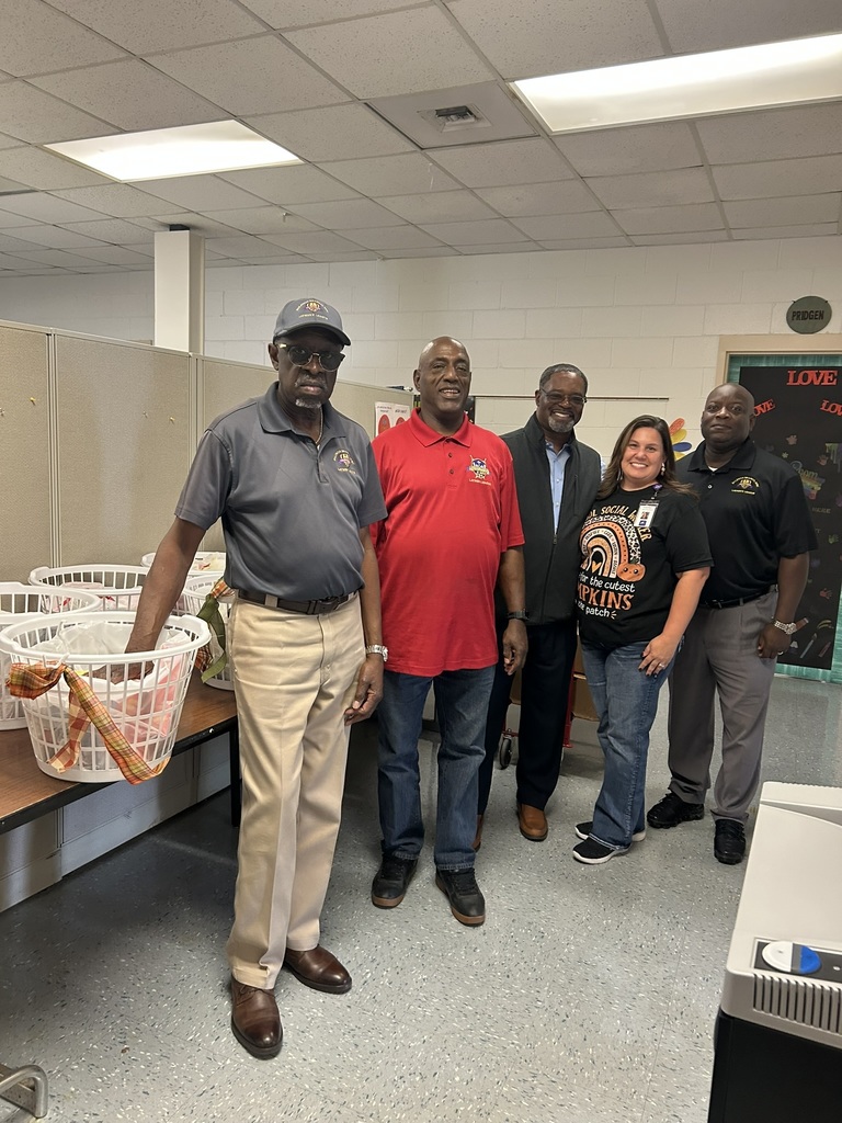 A group of five adults stand together smiling in a school room beside several large white laundry baskets filled with Thanksgiving food items. The baskets are decorated with fall-colored ribbon. The individuals appear to be representatives from Baldwin Branch Church delivering turkey and Thanksgiving meal baskets for school families.