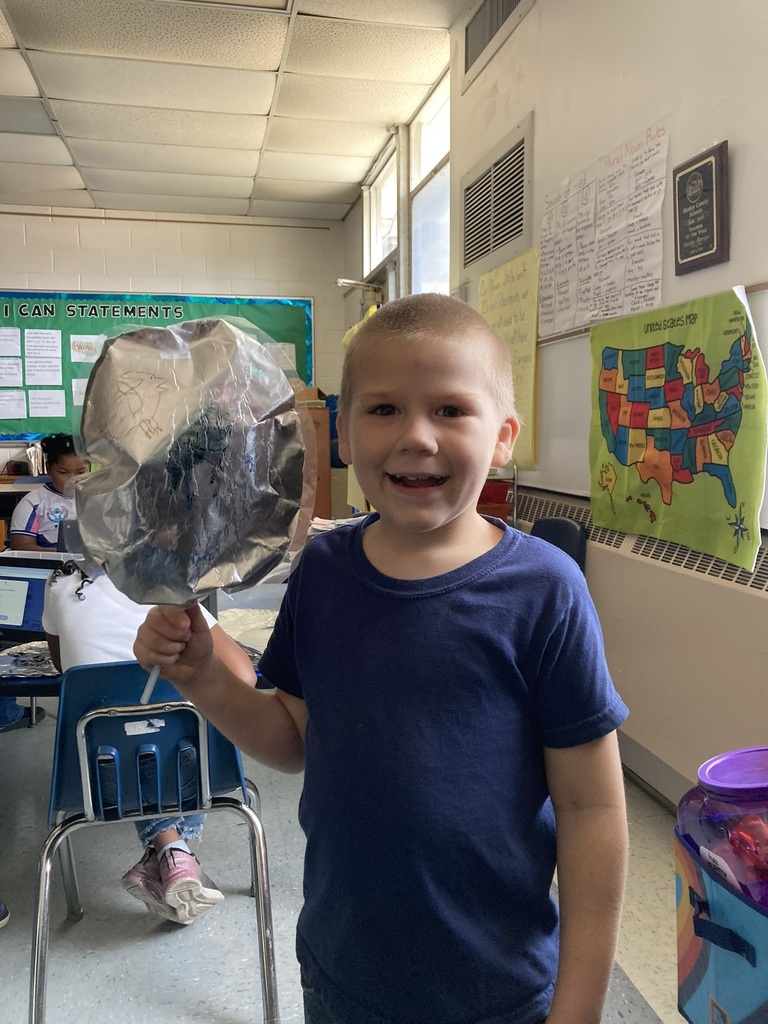 A smiling young student stands in a classroom holding a homemade “parade balloon” made from foil and a straw. His classmates are working at tables in the background.