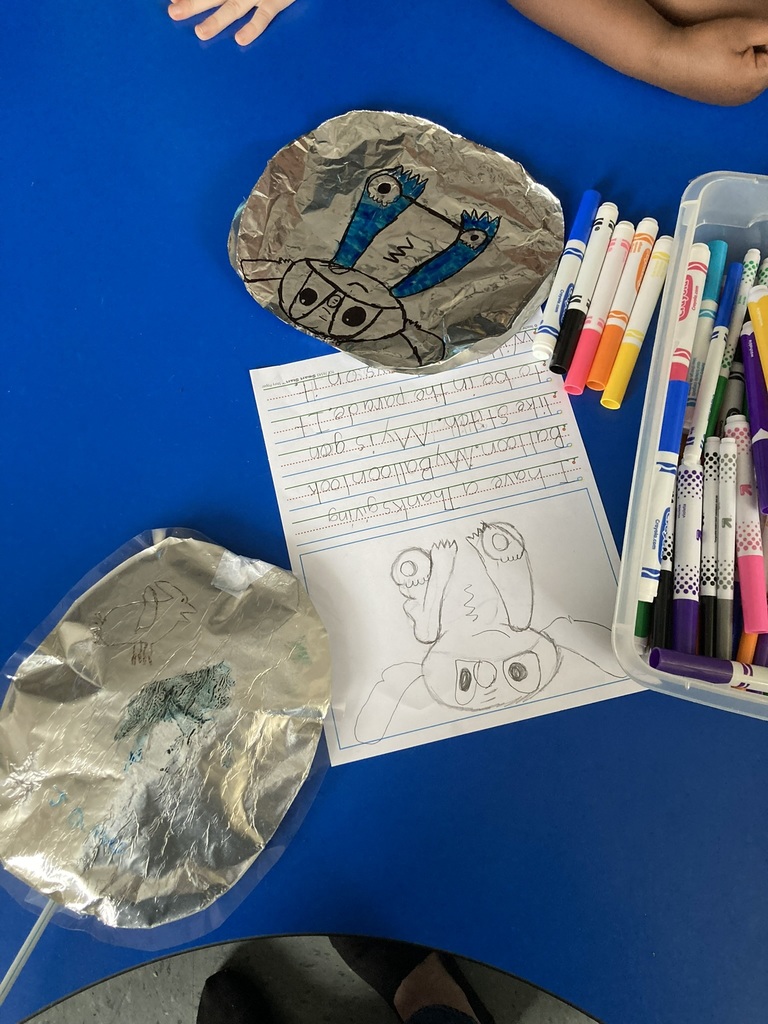 A blue classroom table with foil balloon creations, colorful markers, and a student’s writing and drawing inspired by the book Balloons Over Broadway. One foil balloon features a blue and black character design.