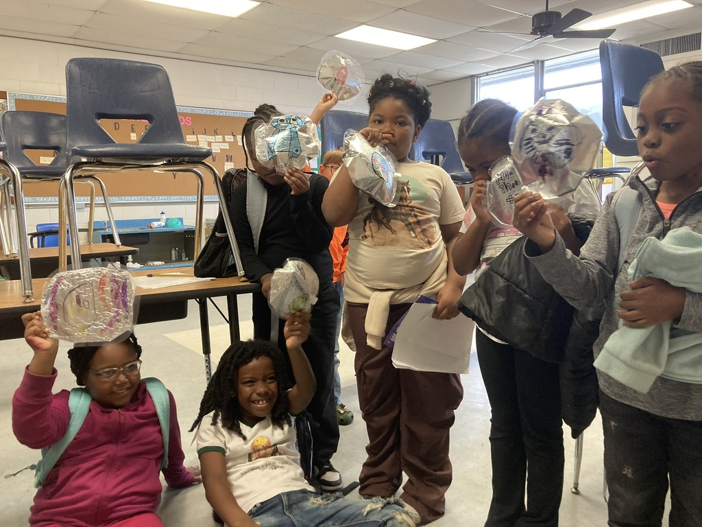 A group of students proudly hold up their homemade foil parade balloons. Some are standing and others sitting on the floor, smiling and showing their creations.