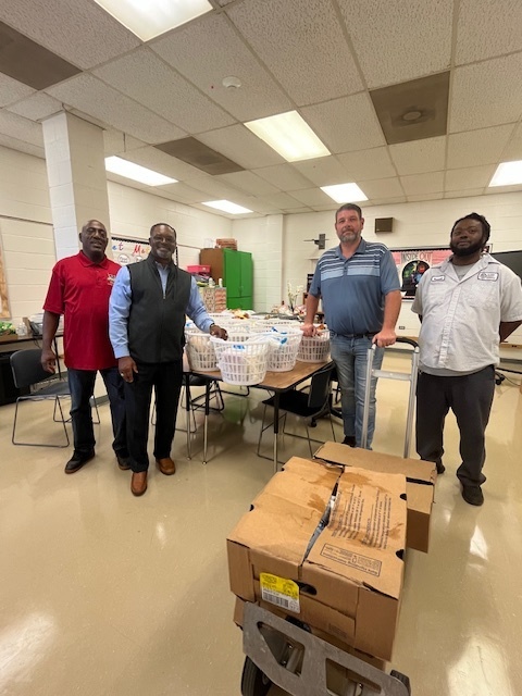 Four men stand together in a school room beside a table filled with baskets and food items. One man stands behind the table while three stand in front of it. A cart with cardboard boxes is positioned on the right side of the image. The room has tiled floors, fluorescent lighting, and shelves and supplies in the background.