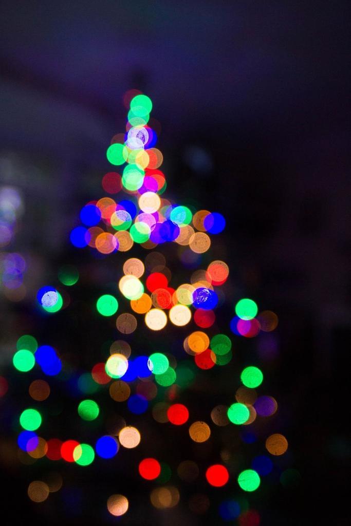 A softly blurred photo of a Christmas tree made of colorful twinkling lights—red, green, blue, yellow, and purple—glowing against a dark background.
