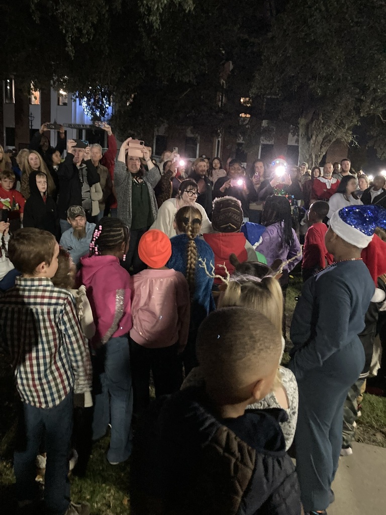 A crowd of children face a large group of adults who are smiling and taking photos with their phones during the Christmas Tree Lighting event. The scene is festive with bright lights and nighttime surroundings.