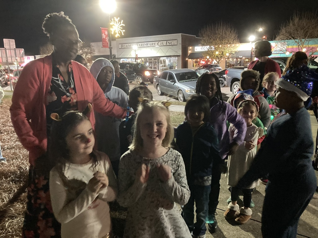 A group of children stand together outside at night, some wearing festive outfits or headbands. An adult in a red sweater stands behind them. Cars, shops, and holiday lights line the street in the background.