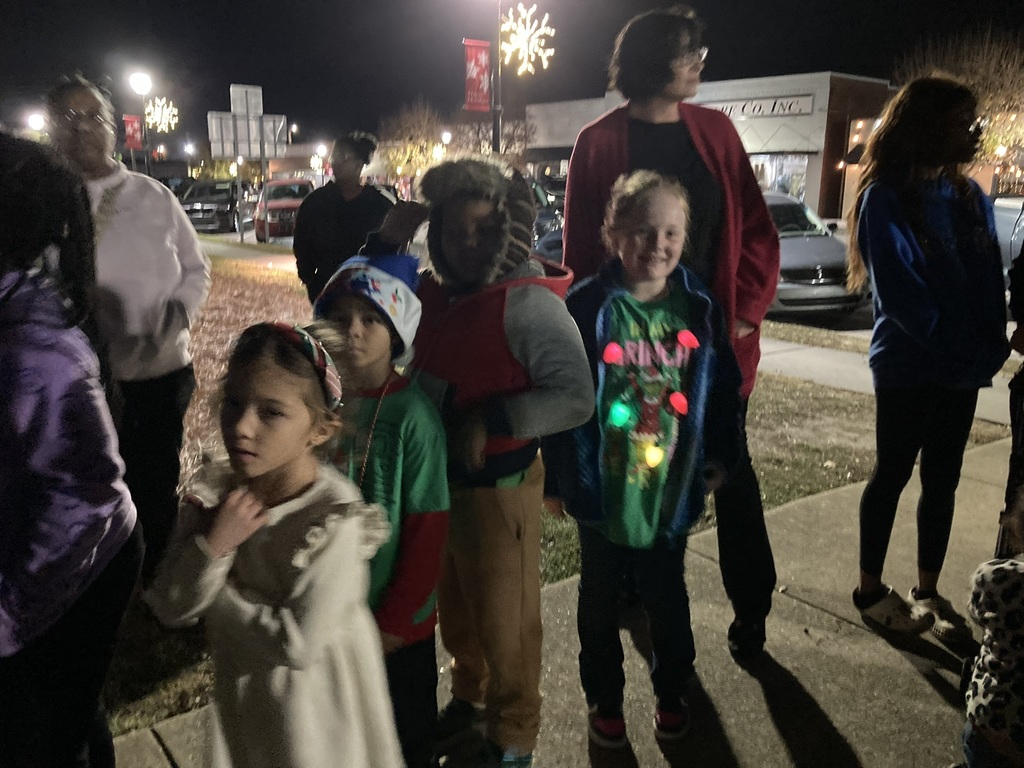Several children stand in a line outdoors at night, wearing colorful jackets and Christmas-themed clothing. A girl in a white dress stands toward the front, and another child wears a hoodie with glowing Christmas lights. Adults stand behind them near the sidewalk.