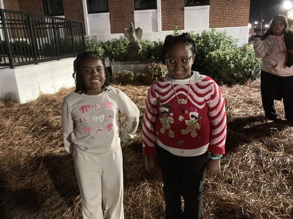 Two young girls stand outside at night on a bed of pine straw. One wears a light-colored outfit with pink bows, and the other wears a red-and-white holiday sweater with gingerbread decorations. Both are smiling with bushes and a brick building behind them.