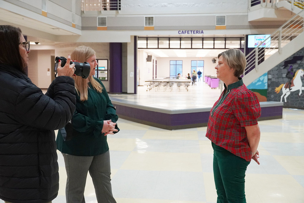 A woman films an interview in the school lobby as another woman in a green blouse speaks with a staff member wearing a red plaid top. The cafeteria is visible in the background.