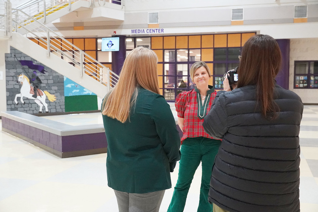 A staff member in a red plaid top smiles while being interviewed by two women, one filming with a camera, in the lobby near the school’s media center.
