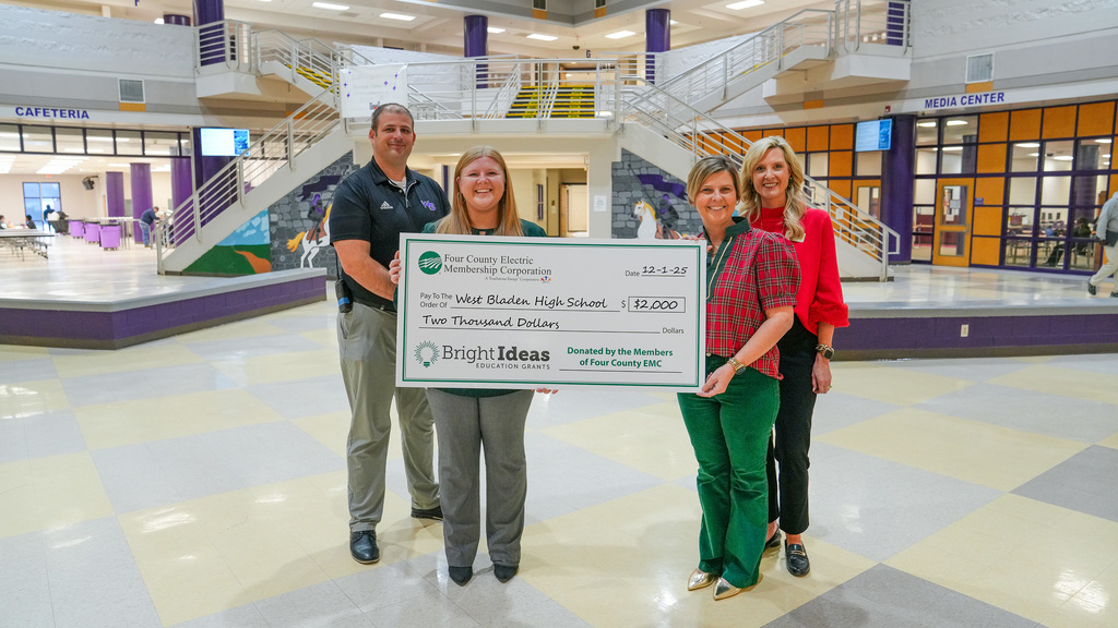 Four adults stand in the West Bladen High School lobby holding an oversized $2,000 Bright Ideas grant check from Four County EMC. The group smiles toward the camera with the cafeteria and media center visible in the background.