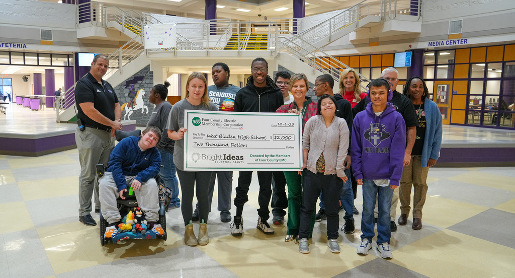 A group of West Bladen High School students and staff stand in the school’s lobby holding an oversized $2,000 Bright Ideas grant check from Four County EMC. A student in a wheelchair is positioned at the front left, and administrators and teachers stand behind the group.