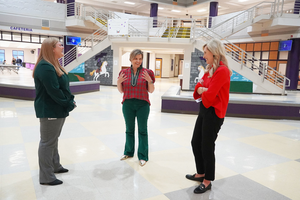 Three women stand in conversation in the school lobby. The woman in red plaid speaks while the others, wearing green and red tops, listen and smile.