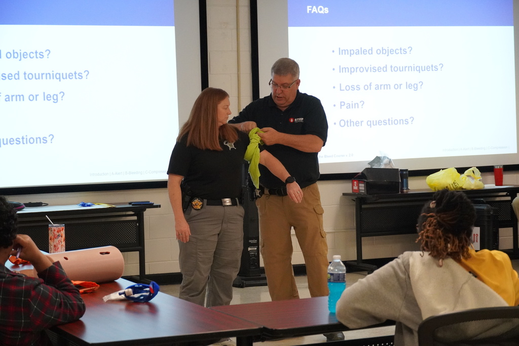 Two instructors stand at the front of the room demonstrating how to apply an improvised tourniquet using a cloth, with a projection screen showing FAQ topics behind them.