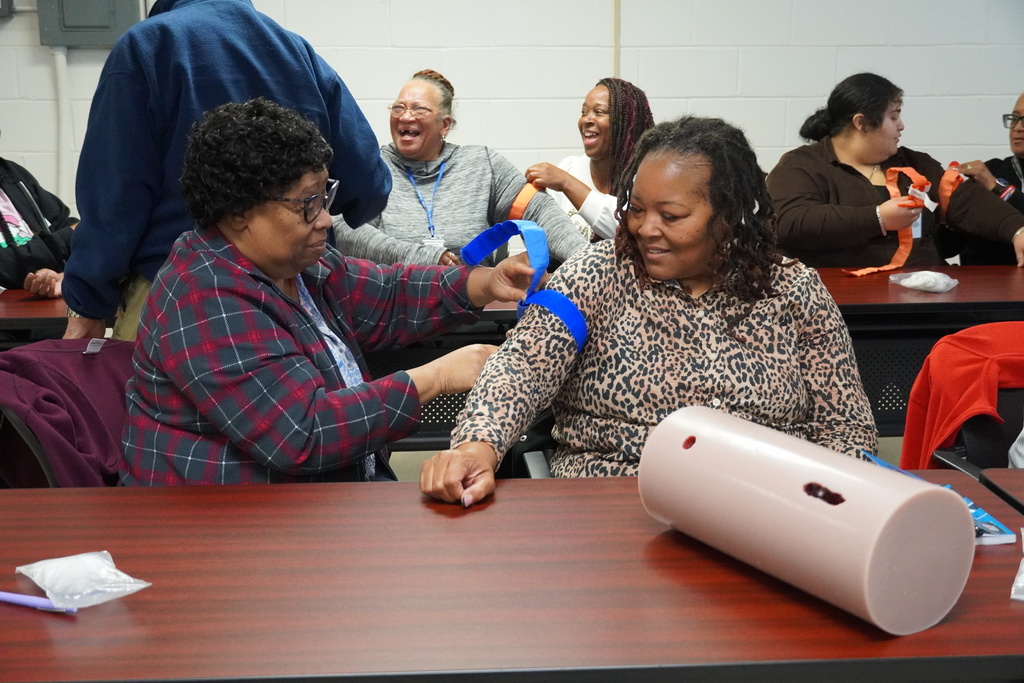 Two women seated at a table practice placing a blue tourniquet on one woman’s arm while others in the room laugh and work on their own training materials.