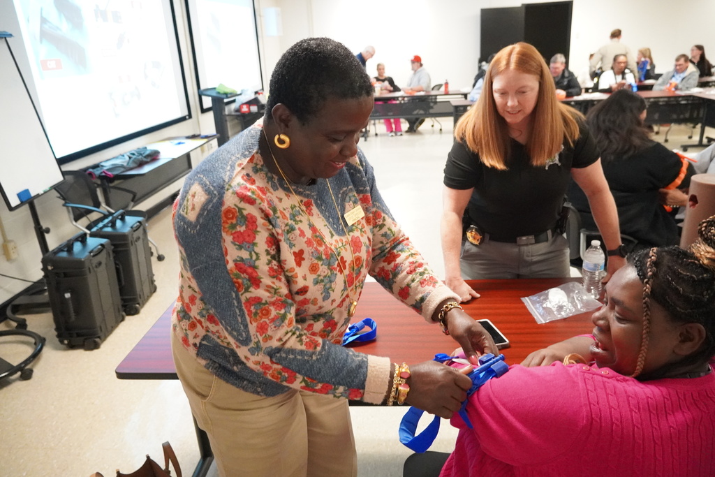 A woman in a floral sweater secures a blue tourniquet on another participant’s arm while a female instructor observes nearby; the room is full of staff practicing the same skill.
