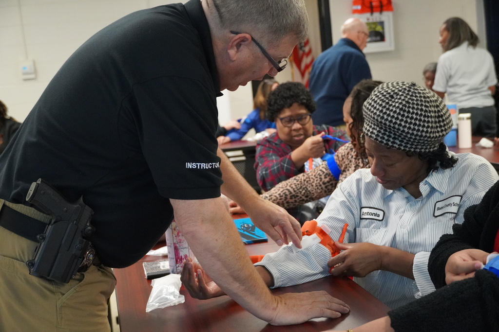 A male instructor leans over to help a woman in a Bladen County Schools uniform apply an orange tourniquet to her arm as participants around them continue practicing.