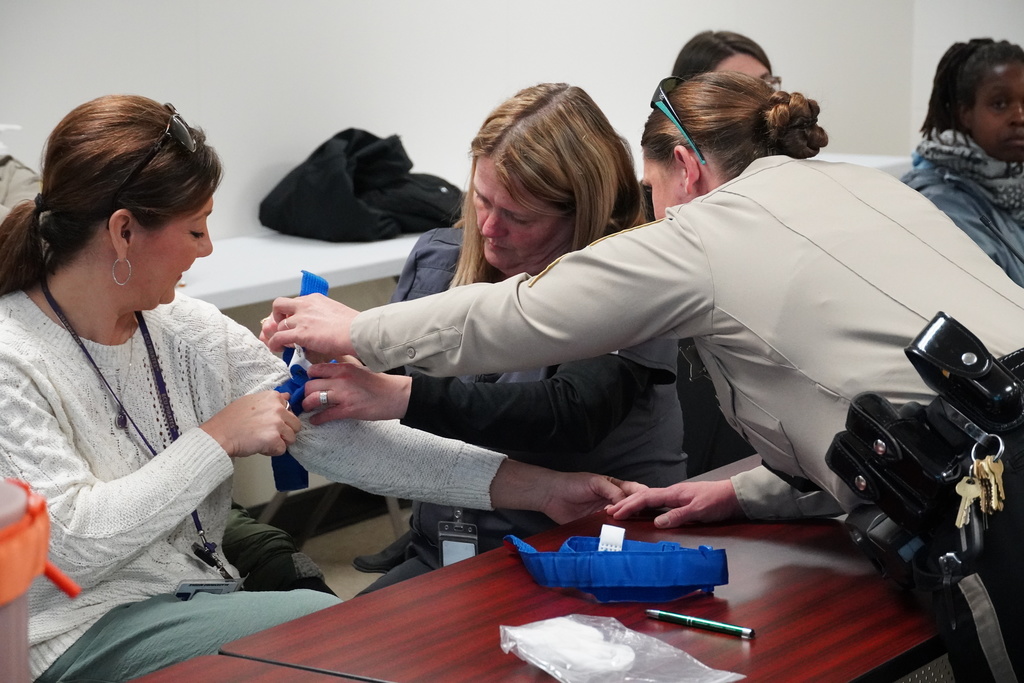 A uniformed officer and another participant assist a woman in tightening a blue tourniquet on her arm during the hands-on training session.