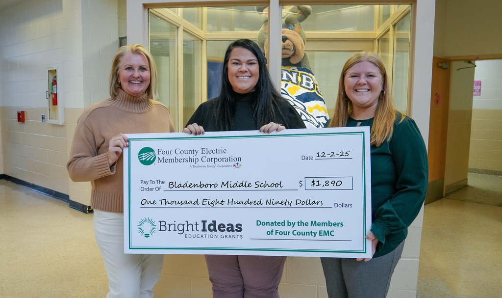 Three women stand in a school hallway holding an oversized ceremonial check made out to Bladenboro Middle School for $1,890 from Four County Electric Membership Corporation’s Bright Ideas Education Grants program; the school’s bear mascot is visible behind them through a glass window.
