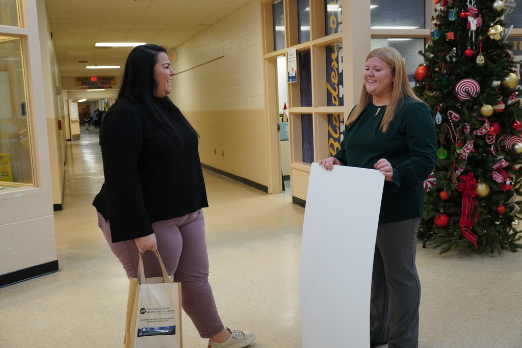 Two women stand in a school hallway smiling and talking; one holds a tote bag and the other holds a blank poster board. A decorated Christmas tree is visible behind them.