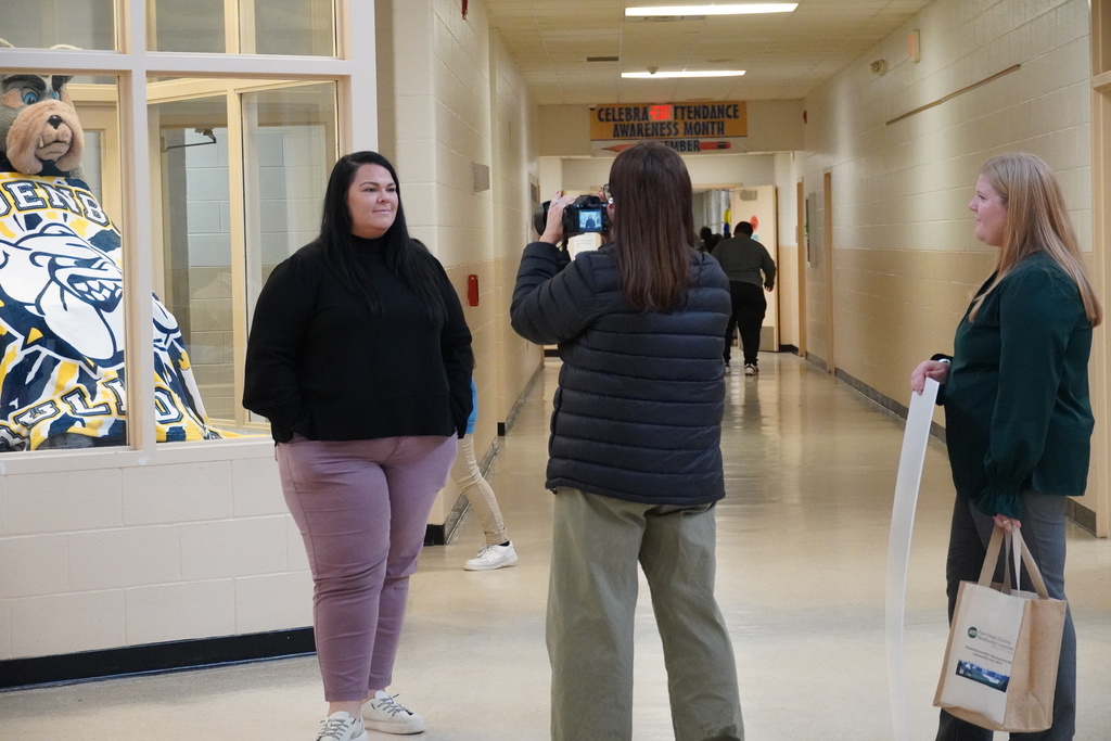 A woman stands in a school hallway as another woman photographs her, while a third woman holding a tote bag and poster board looks on. The school’s bear mascot costume is visible through a window behind them.