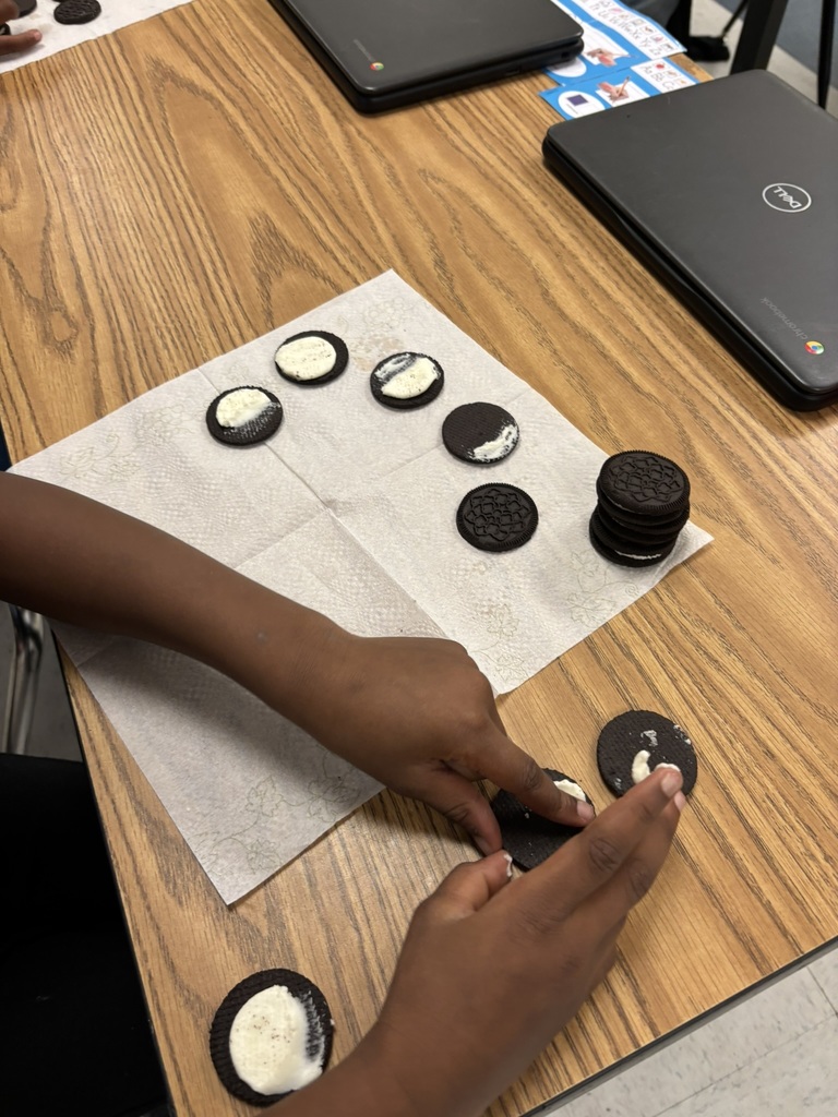 A student’s hands pressing Oreo halves together while creating moon phases with the cream filling. Several completed moon phase cookies are arranged on a napkin nearby, next to Chromebooks on the table.
