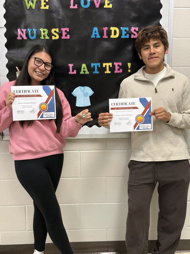 Two smiling students hold HOSA participation certificates in front of a "We Love Nurse Aides a Latte" bulletin board.