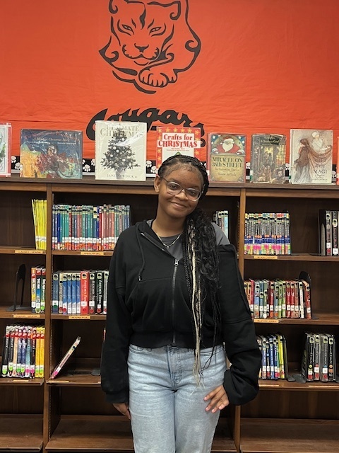A student stands smiling in front of a library bookshelf at Elizabethtown Middle School. She is wearing glasses, a black jacket, and light blue jeans. Behind her are shelves filled with colorful books and a bright orange wall featuring the Cougar mascot logo. Several displayed books are arranged along the top of the shelf.
