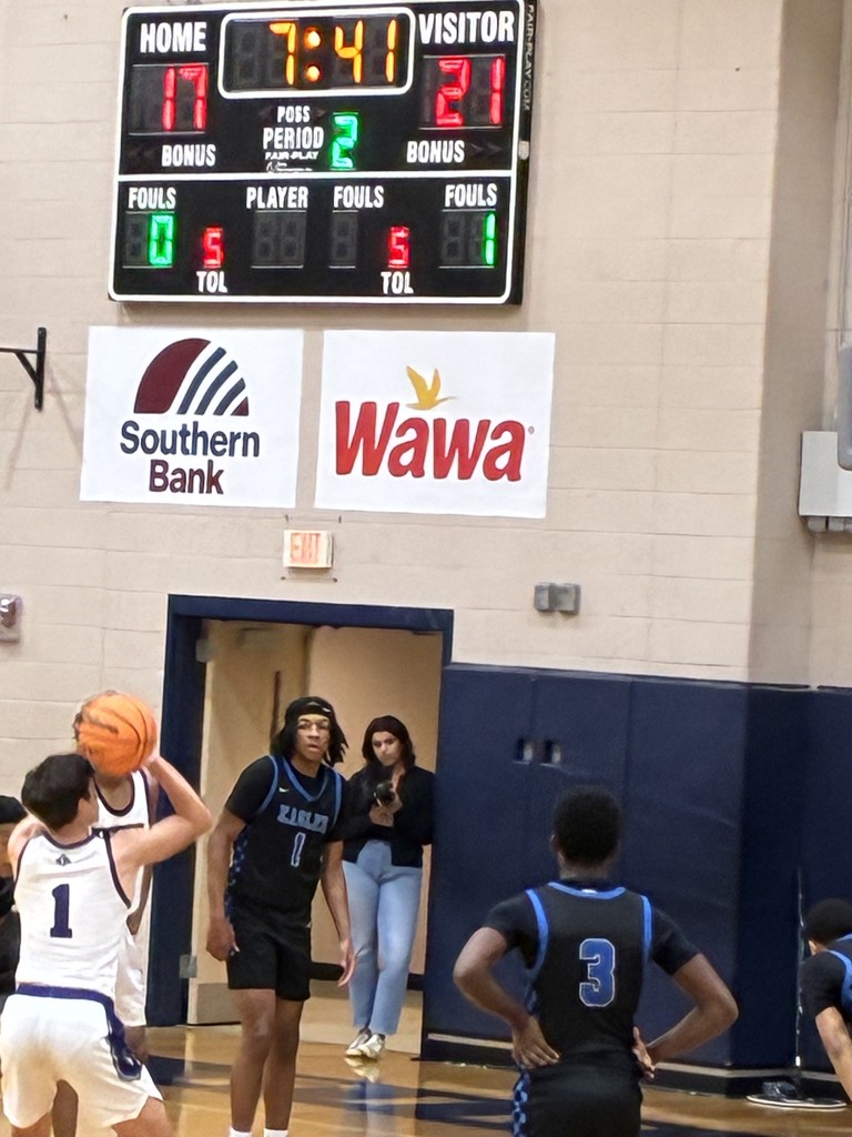 A basketball team in black and purple uniforms warms up in a gym with "NIGHTHAWKS" banners and a scoreboard in the background.