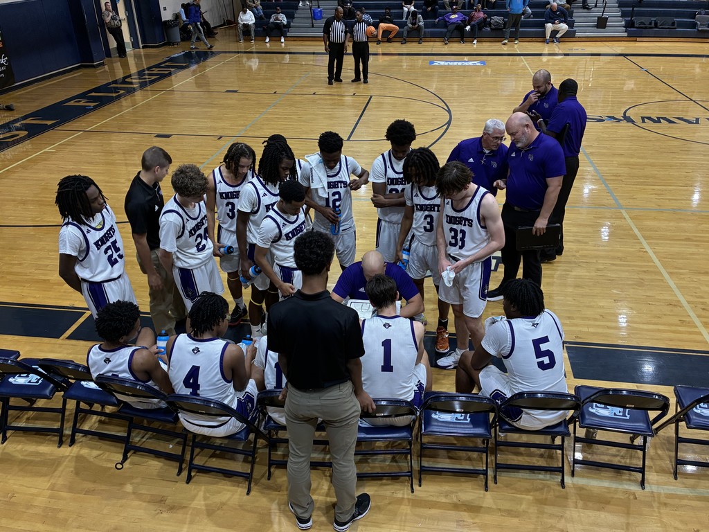 Coach with whiteboard addresses huddling basketball team in white uniforms during a game.