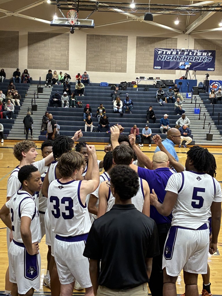 A high school basketball team in white and purple uniforms huddles on the court with hands raised, spectators in bleachers.
