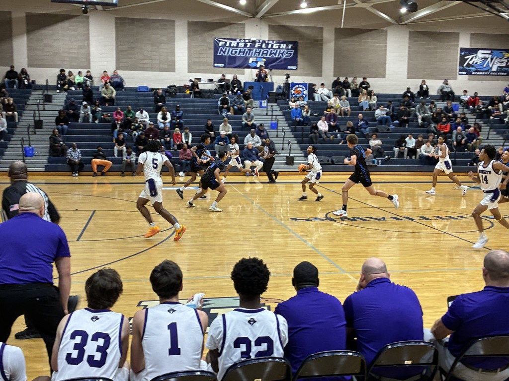 Basketball game in progress between white/purple and black/blue teams, with players, coaches, and spectators.