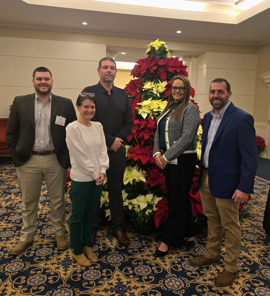 Group of five adults standing indoors in front of a large red, white, and yellow poinsettia Christmas tree display, smiling for a posed photo.
