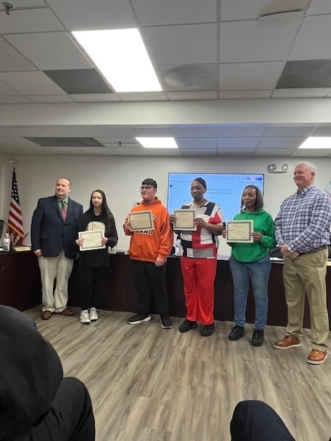 A group of seven people stand at the front of a meeting room during a Bladen County Schools Board Meeting. Four individuals in the center—two students and two staff members—hold certificates of recognition. One student wears an orange Elizabethtown Middle School hoodie, and the other wears a red-and-white jacket. The two staff members stand behind them holding certificates. Two school officials stand at each end of the group. An American flag and a presentation screen are visible in the background.