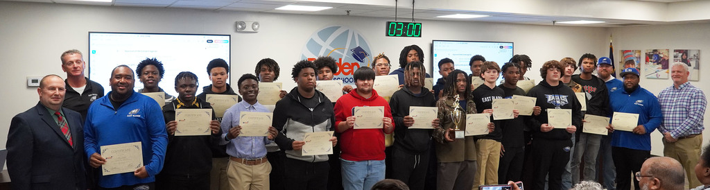 A large group of student athletes and coaches stands in a boardroom holding certificates, with one student also holding a trophy. They pose for a recognition photo, with meeting screens and district branding visible behind them.