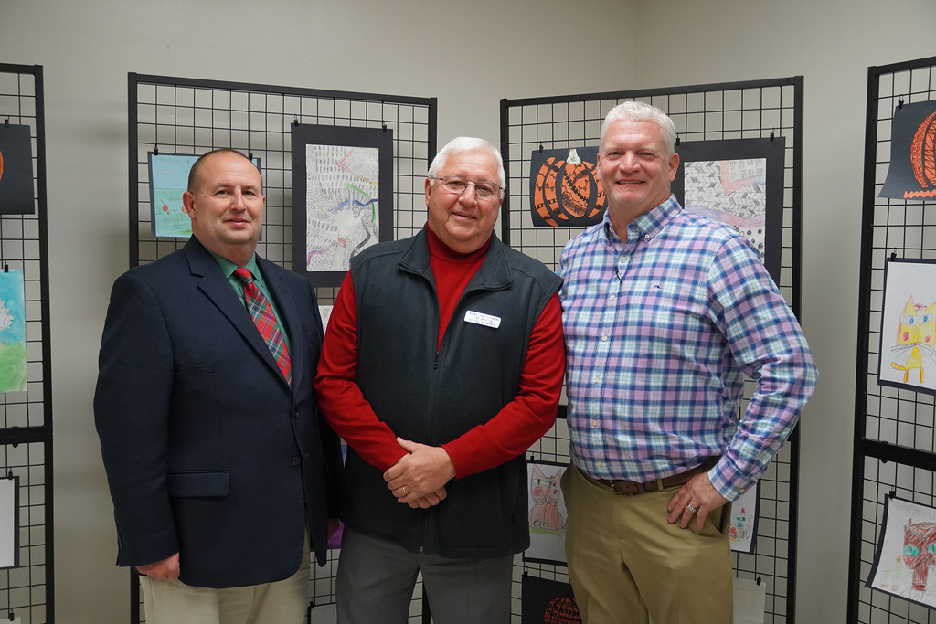 Three district leaders stand together and smile for a photo in front of student artwork displayed on grid panels.