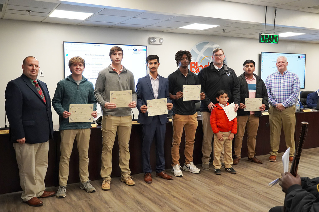 A group of students stands in a boardroom holding certificates, joined by two adults and a young child. They face the camera for a recognition photo, with meeting screens and nameplates visible in the background.