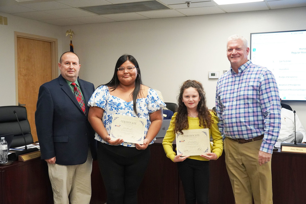Two students hold certificates while standing between two adults in a boardroom. One student wears a white and blue floral top, the other a yellow sweater. The men on each side wear a suit and a plaid shirt, and all look toward the camera.