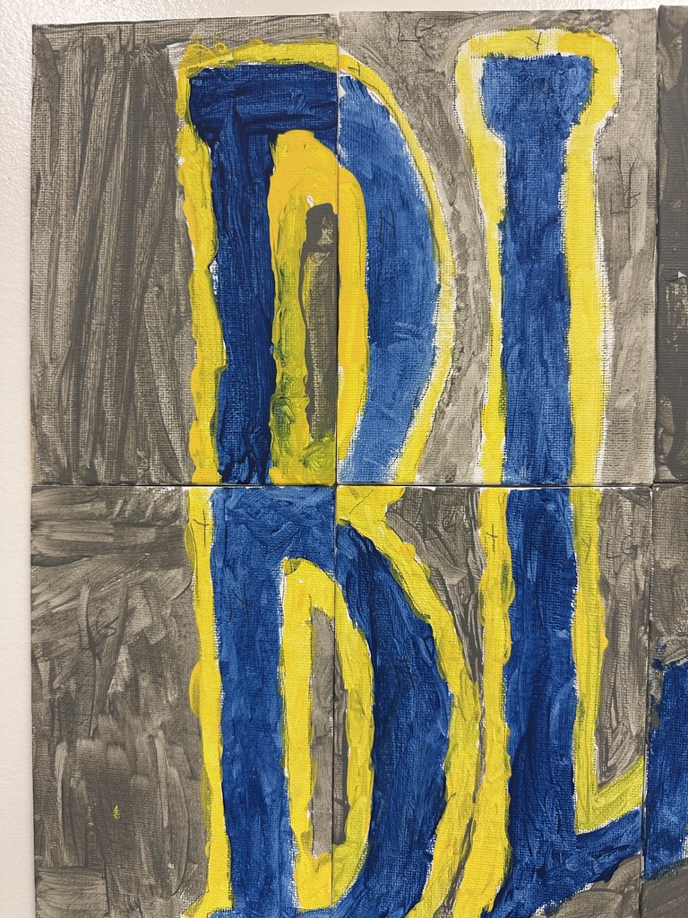 Close-up of a painted blue and yellow letter ‘B’ on a gray textured background, created on small square canvas tiles.