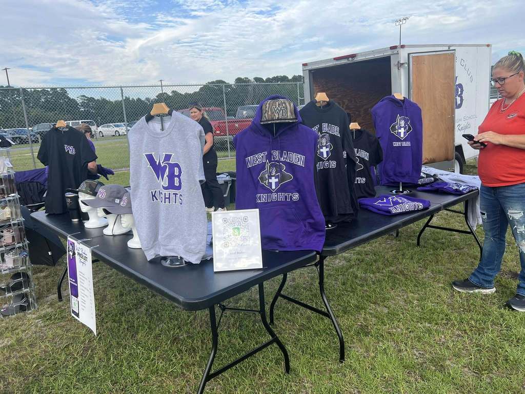 West Bladen Knights merchandise stall with purple and black hoodies, shirts, and hats displayed on outdoor tables.