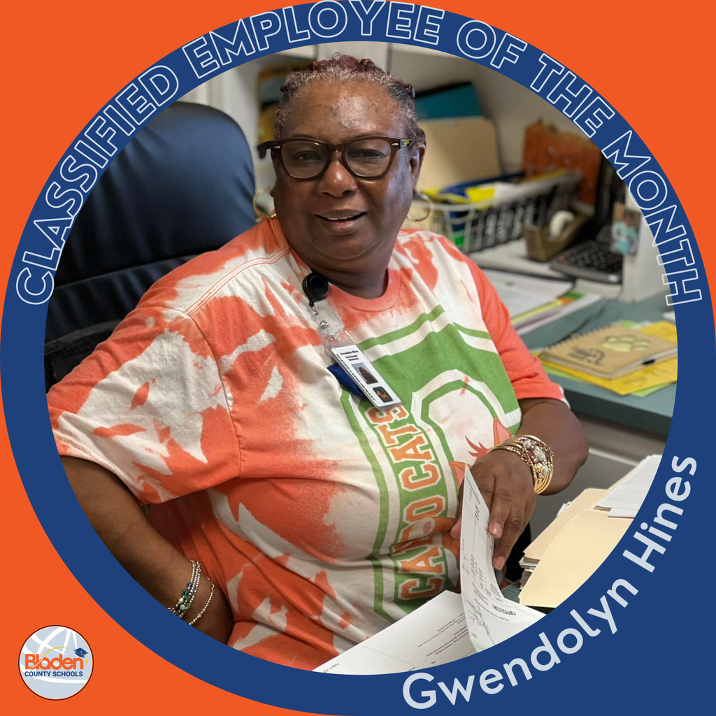 A woman wearing glasses and an orange-and-white tie-dye T-shirt sits at a desk holding paperwork. The circular frame around the photo reads ‘Classified Employee of the Month – Gwendolyn Hines.