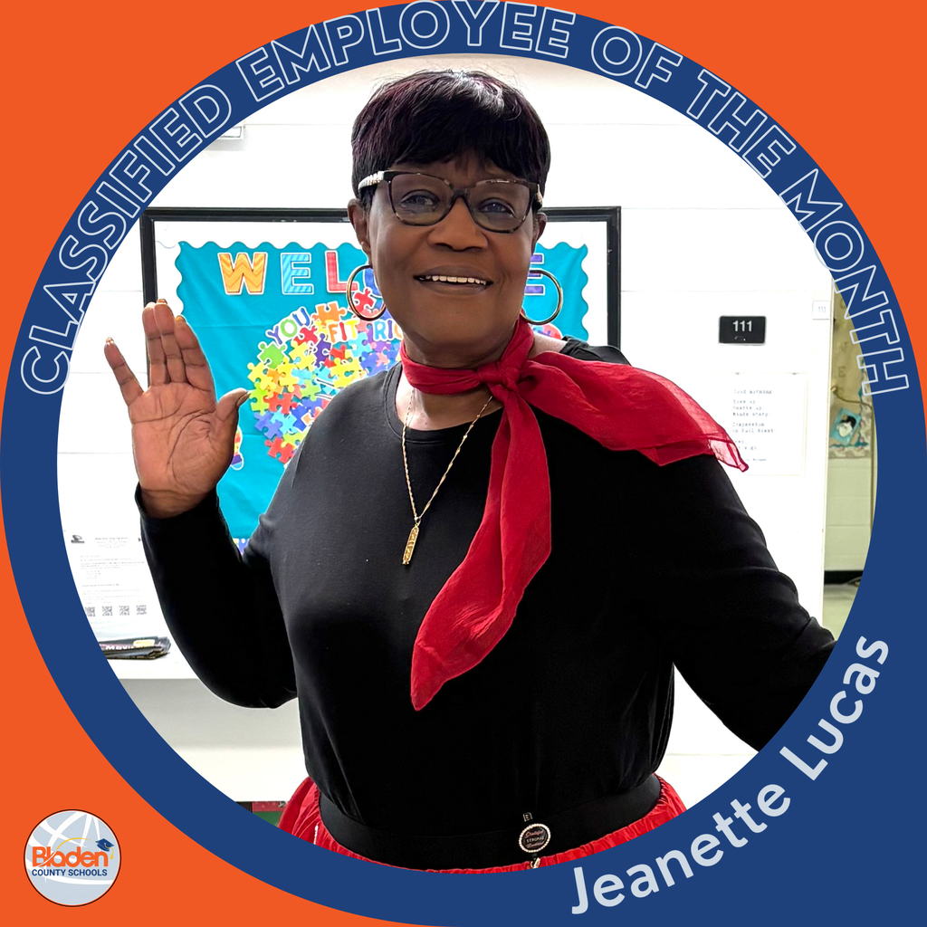 A woman wearing glasses, a black top, and a red scarf smiles and raises her hand while standing in a school hallway. The circular frame reads ‘Classified Employee of the Month – Jeanette Lucas.