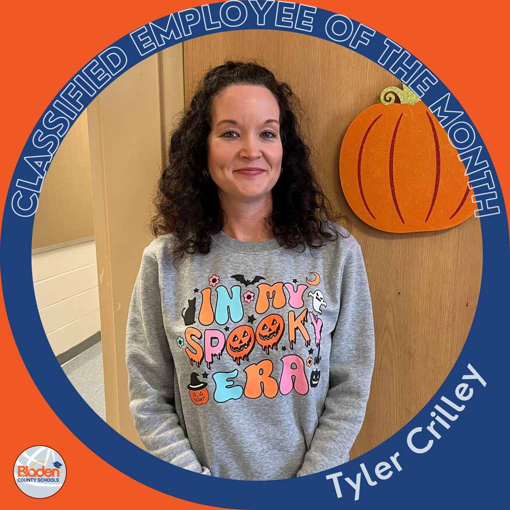 A woman with curly dark hair stands in a hallway beside a decorative pumpkin cutout. She wears a gray sweatshirt that says “In My Spooky Era” in colorful letters. A circular orange-and-blue frame reads “Classified Employee of the Month,” with “Tyler Crilley” at the bottom and the Bladen County Schools logo in the corner.
