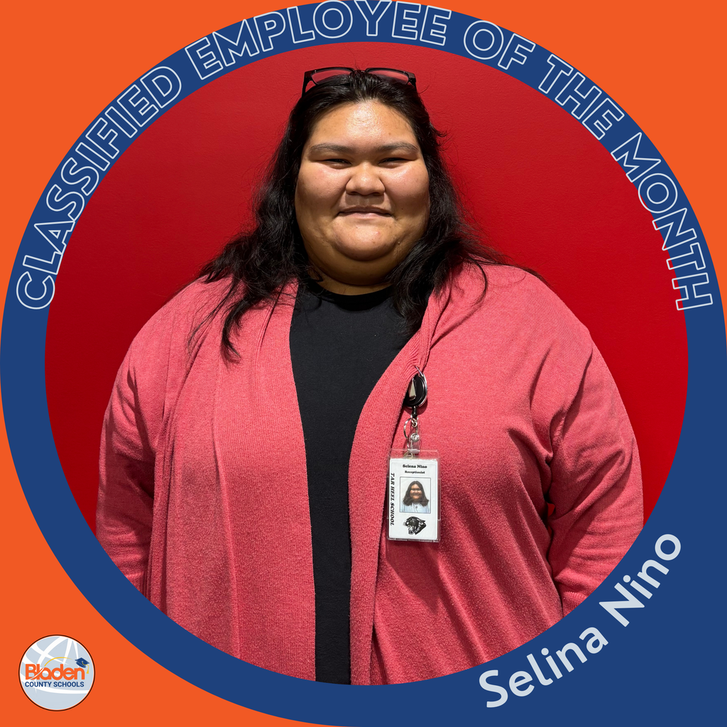 A woman stands in front of a bright red wall wearing a black shirt and a pink cardigan. Her ID badge hangs from a lanyard. A circular orange-and-blue frame reads “Classified Employee of the Month,” with “Selina Nino” at the bottom and the Bladen County Schools logo in the corner.