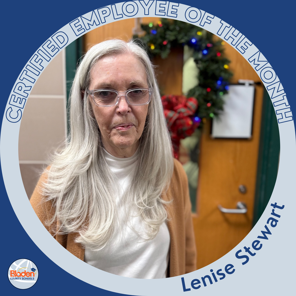 A woman with long silver hair and glasses stands indoors near a door decorated with a holiday wreath. She is wearing a tan cardigan over a white turtleneck. The photo is bordered by a blue-and-gray circular “Certified Employee of the Month” frame labeled “Lenise Stewart
