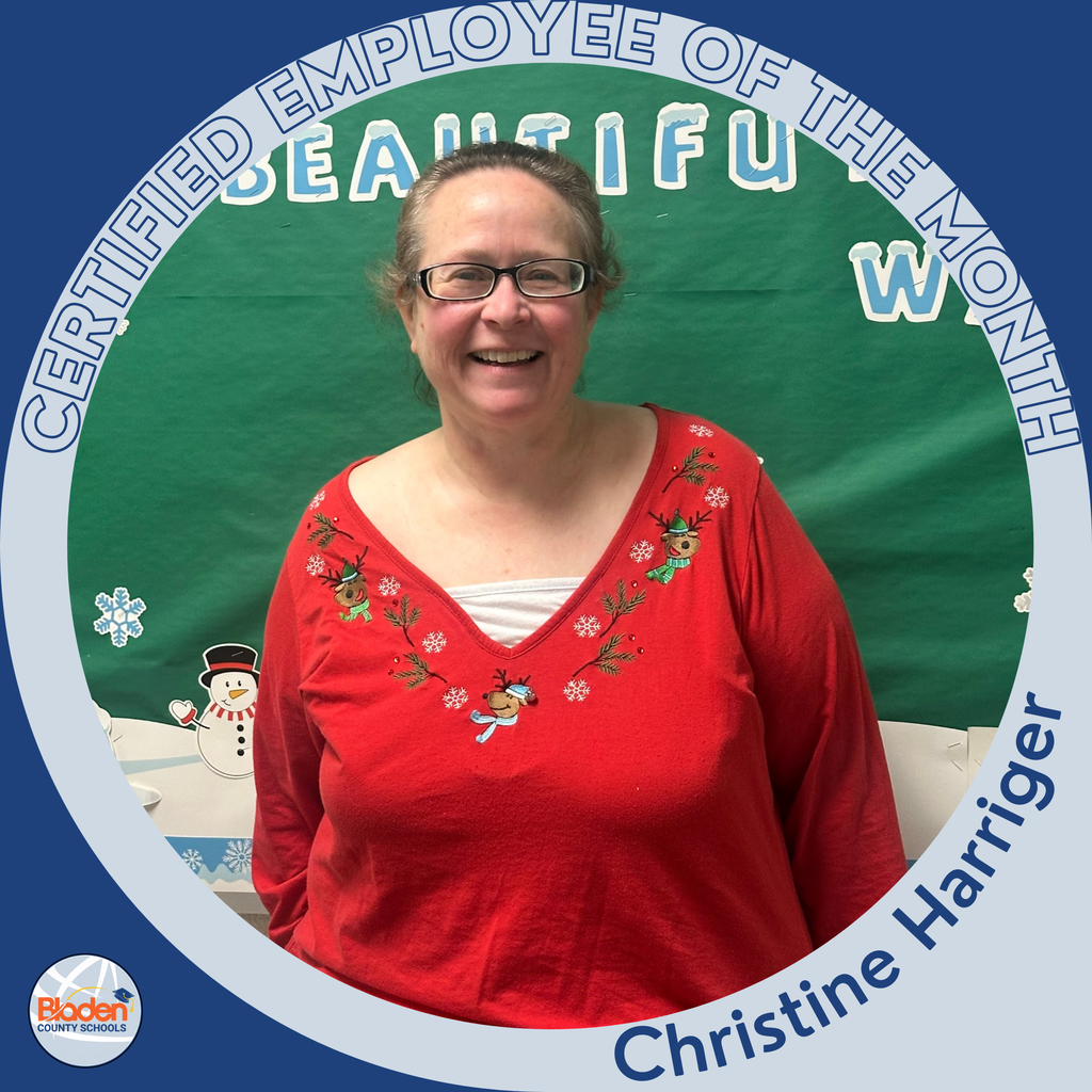 A smiling woman wearing glasses and a red holiday-themed top with embroidered reindeer stands in front of a green bulletin board decorated with winter-themed cutouts. A circular blue-and-gray “Certified Employee of the Month” frame surrounds the image, with the name “Christine Harriger” at the bottom